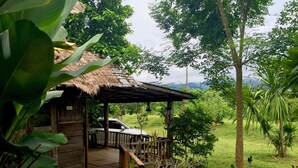 A wooden cabin with a thatched roof, a car parked outside, and a grassy area with trees and chickens. - Sipsongpanna Resort Pai (Pai)