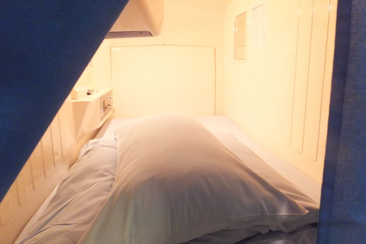 A narrow, single-bed room with a headboard, a small bedside table, and a wall-mounted light.