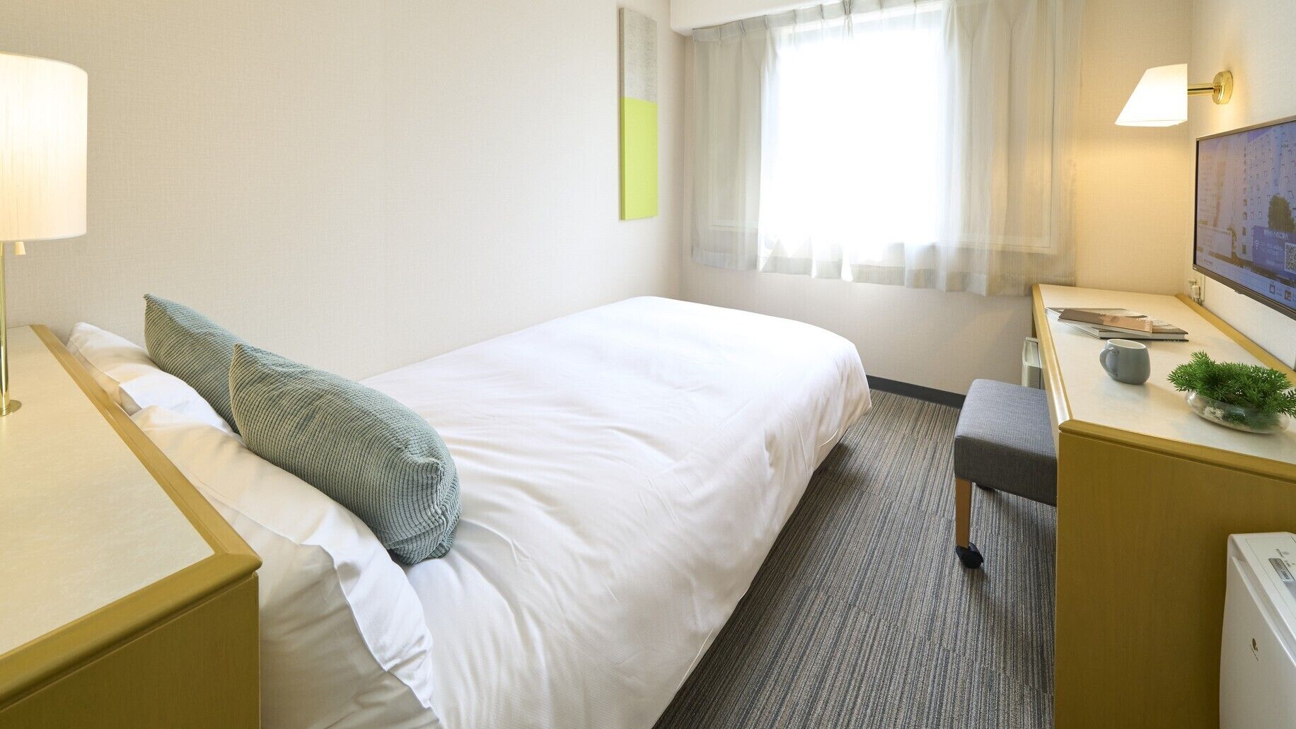 A hotel room with a bed, bedside tables, a desk with a chair, a small plant, and a window with curtains.