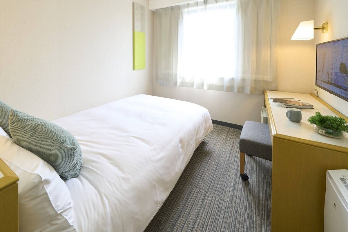 A hotel room with a bed, bedside tables, a desk with a chair, a small plant, and a window with curtains.