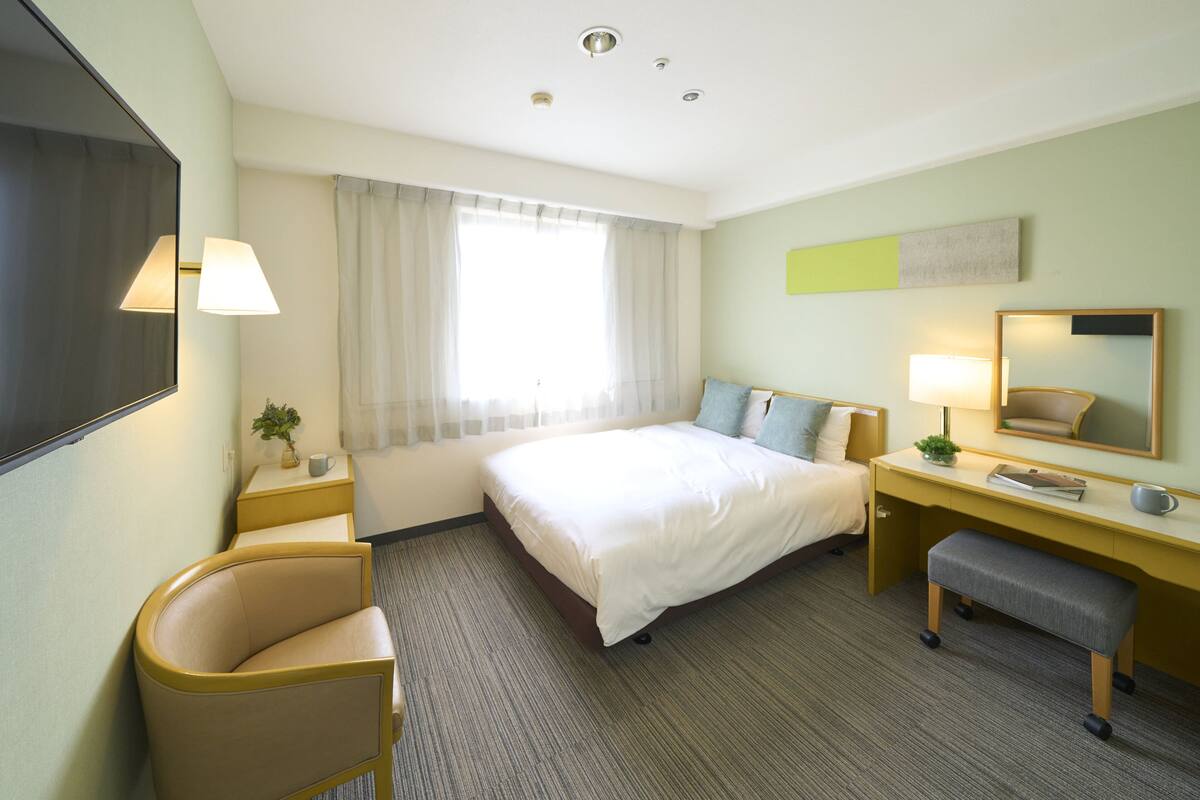 A hotel room with a bed, desk, chair, and a large window with curtains.