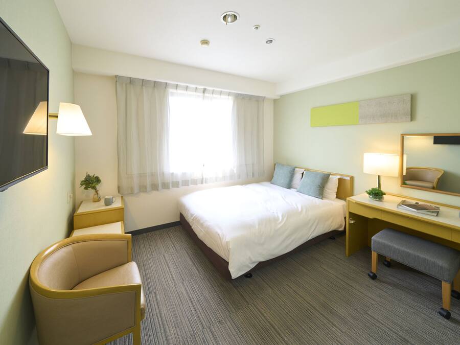 A hotel room with a bed, desk, chair, and a large window with curtains.