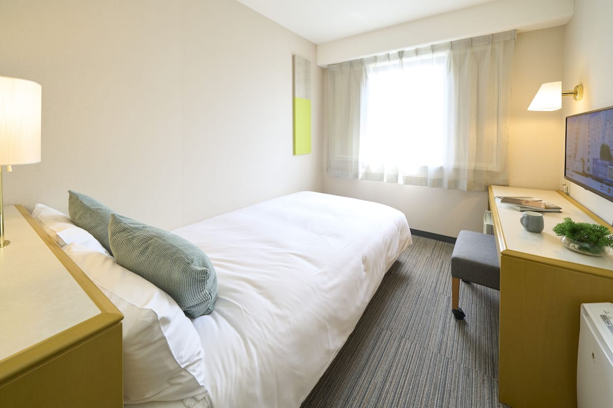 A hotel room with a bed, bedside tables, a desk, a chair, a television, and a window with curtains.
