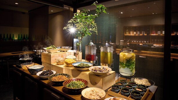 A buffet setup with various dishes and drinks, a decorative plant, and a bar area with bottles in the background. - Hotel Resol Machida (Machida)