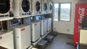 A laundromat with multiple washing machines, a Coca-Cola refrigerator, and a window providing a view of the city. - Hotel Resol Machida (Machida)