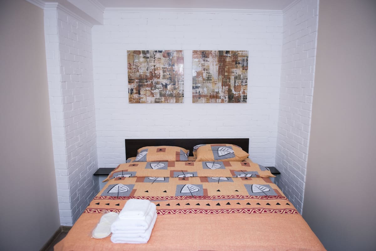 Une chambre avec un lit, des murs en briques blanches, deux tableaux et une couvre-lit à motifs.