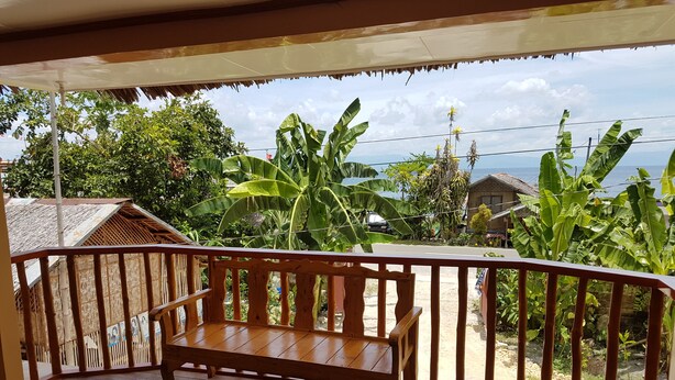 A wooden bench on a porch with a view of a tropical landscape, including banana trees and a body of water. - Lucci Pension House (Alegria)