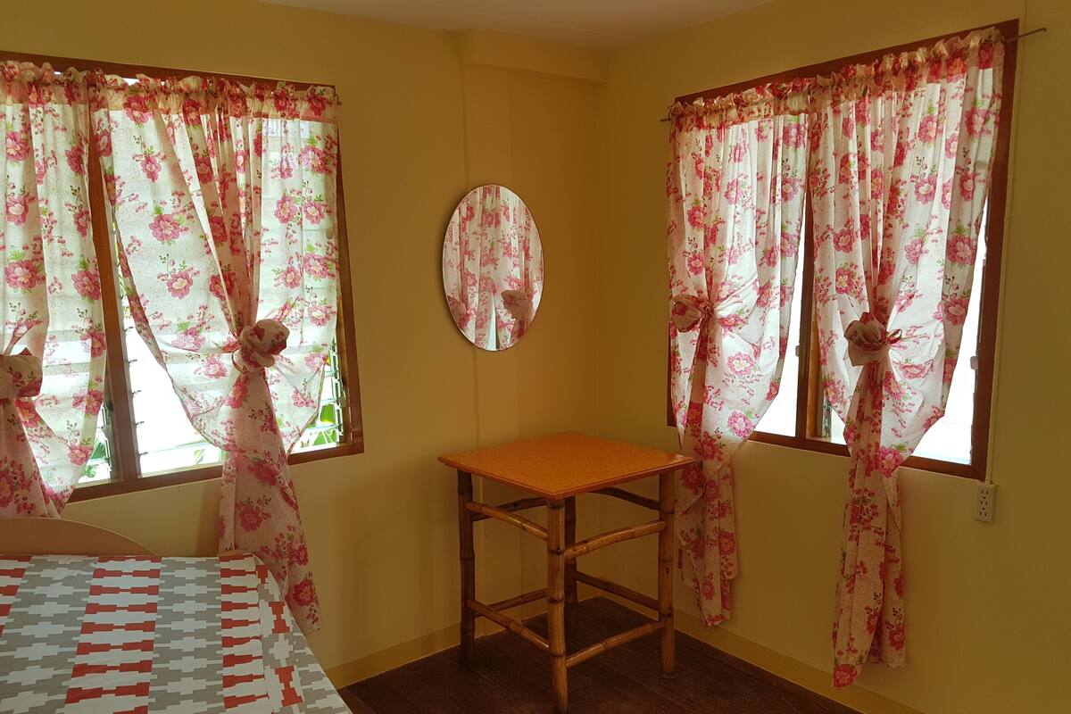 A room with two windows, a wooden chair, a mirror, and a bed with a patterned bedspread.