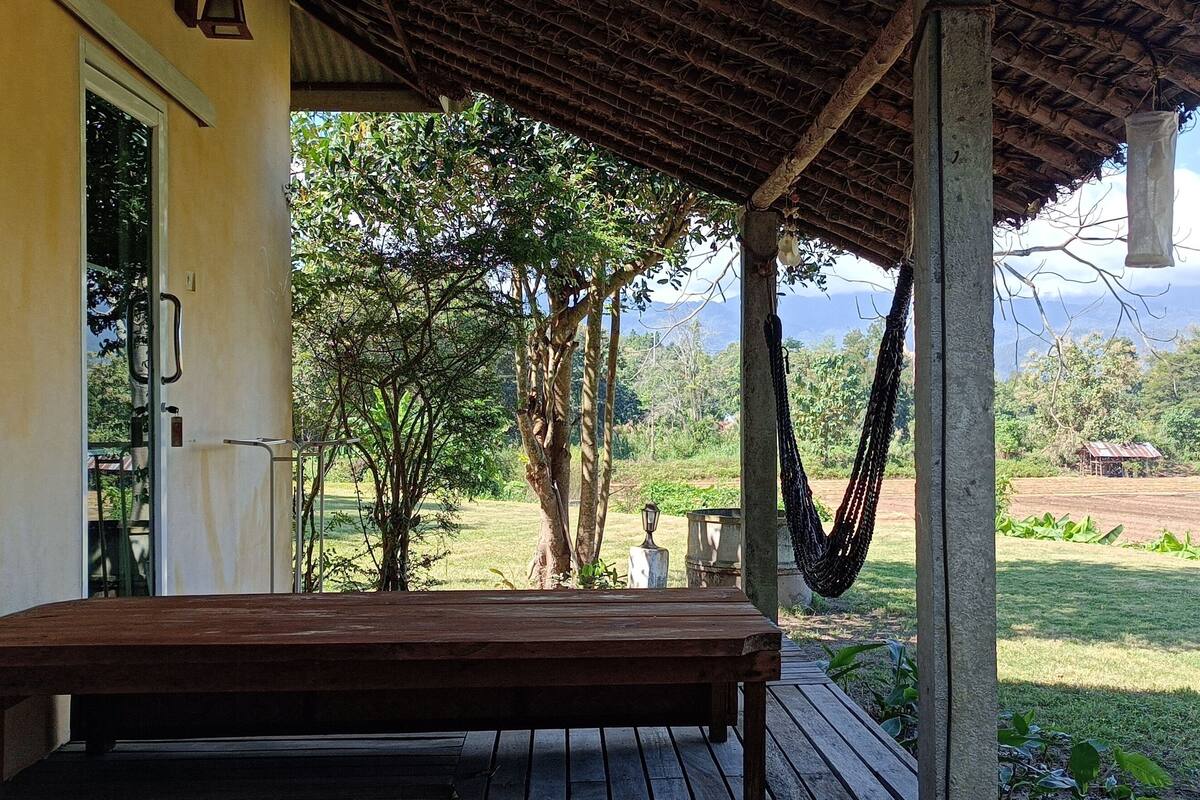 A wooden bench under a covered porch with a view of a green field and mountains in the distance.