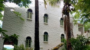 Um edifício branco com várias janelas em arco e um jardim exuberante, com diversas plantas e uma palmeira.