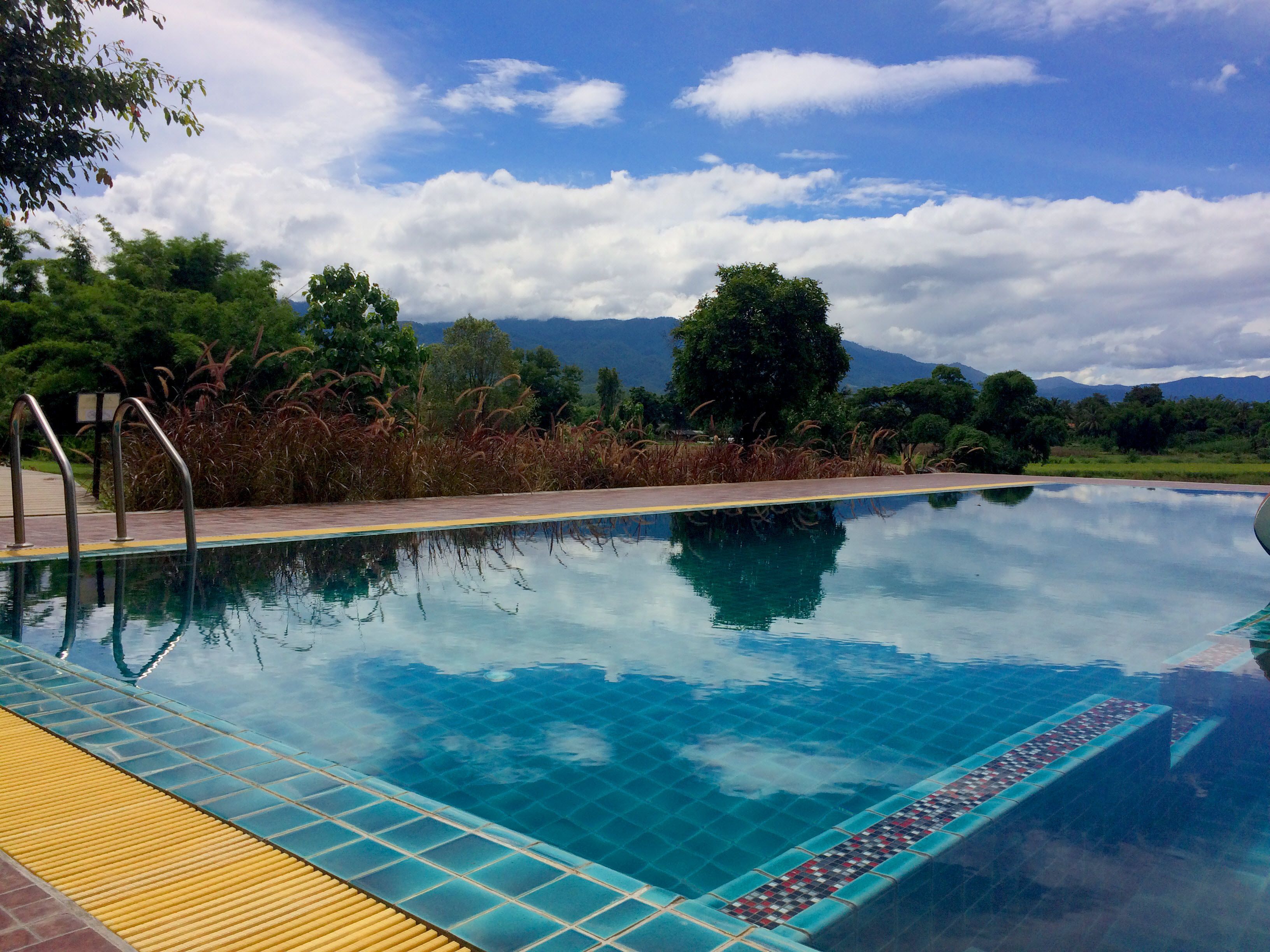 A clear swimming pool with a tiled edge, surrounded by greenery and mountains in the background.