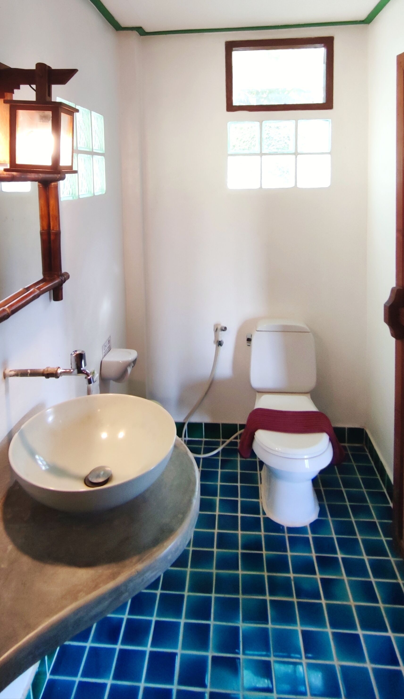 A bathroom with a blue-tiled floor, a white sink with a wooden basin, a toilet with a red cloth, and a window with a wooden frame.
