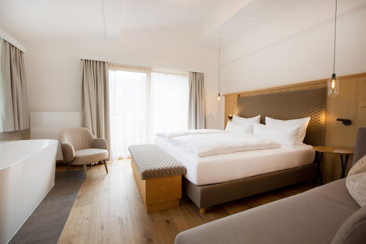 A modern hotel room with a large bed, a sitting area with a bench, a small table, and a bathtub.