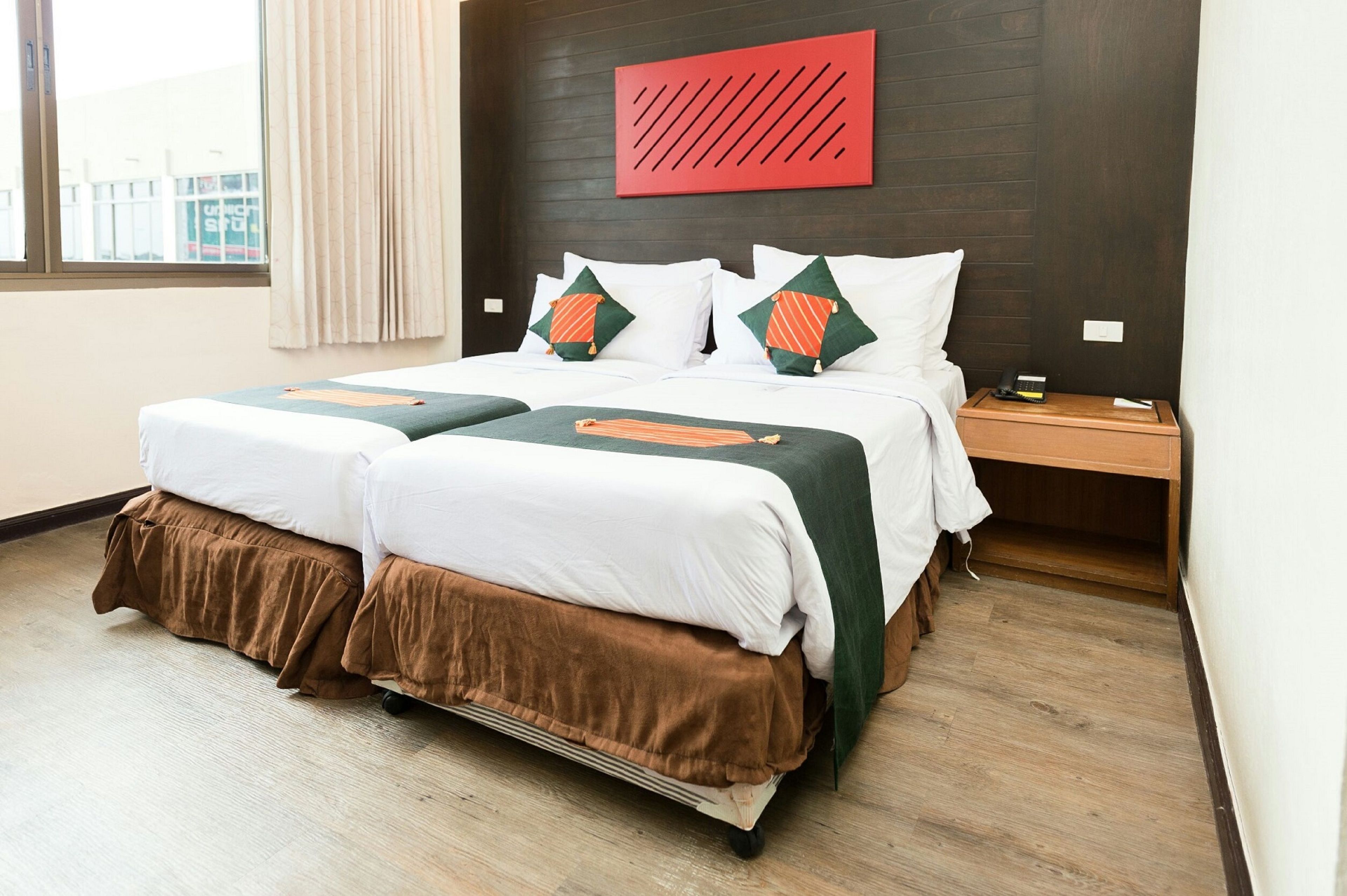 A double bed with a brown skirted base, white linens, and two decorative pillows. A red abstract artwork hangs above the bed.
