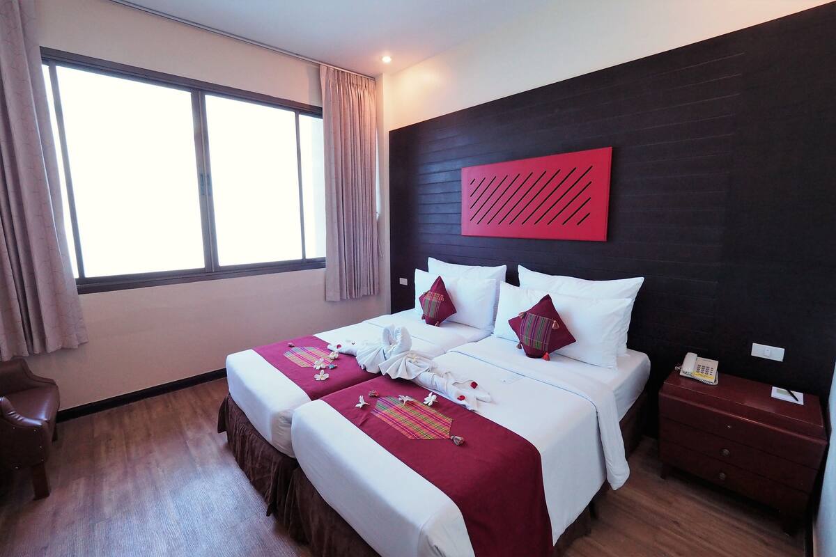 A hotel room with two beds, a large window, a wooden floor, and a red accent wall.
