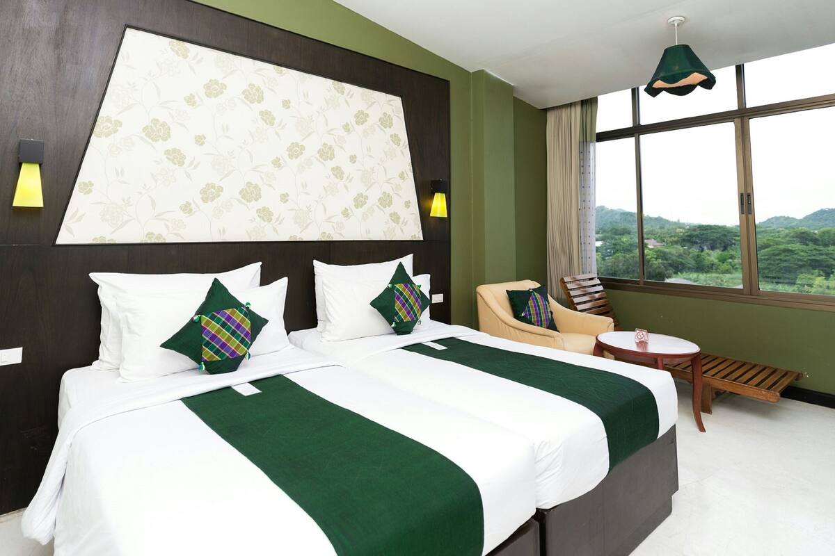 A hotel room with a large bed, two pillows, a chair, a small table, and a view of greenery through the window.