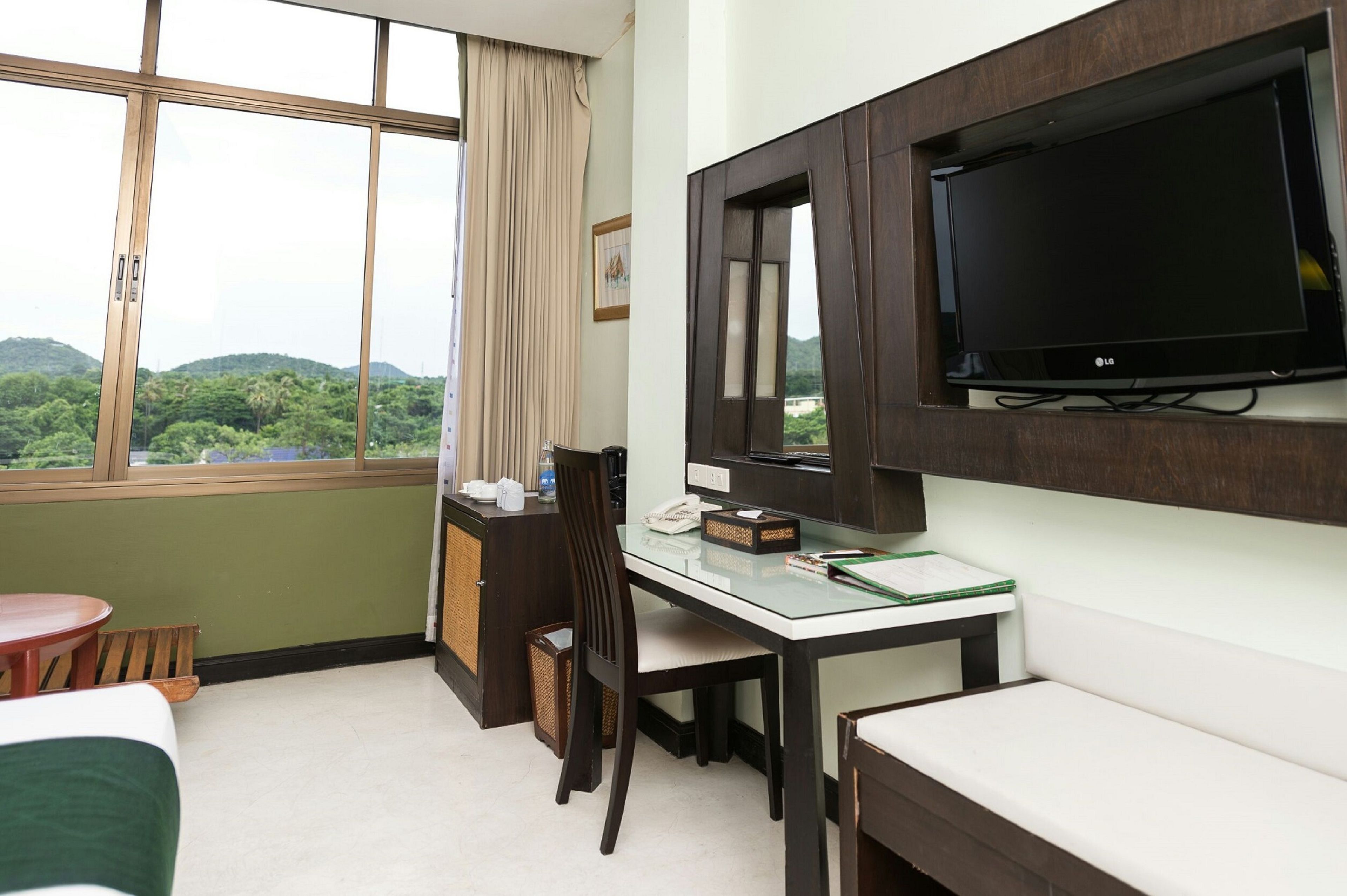 A hotel room with a desk, chair, TV, and a view of greenery through the window.