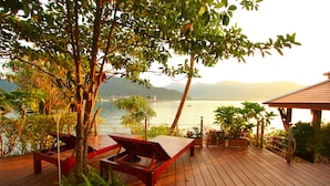 A wooden deck with lounge chairs and tables overlooking a body of water with mountains in the background. - Bhuvarin Resort (Ko Chang)
