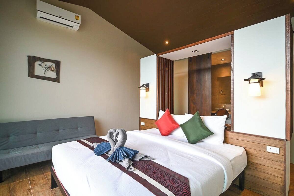 A hotel room with a large bed, a grey sofa, a wall-mounted air conditioner, and a mirror reflecting the room.