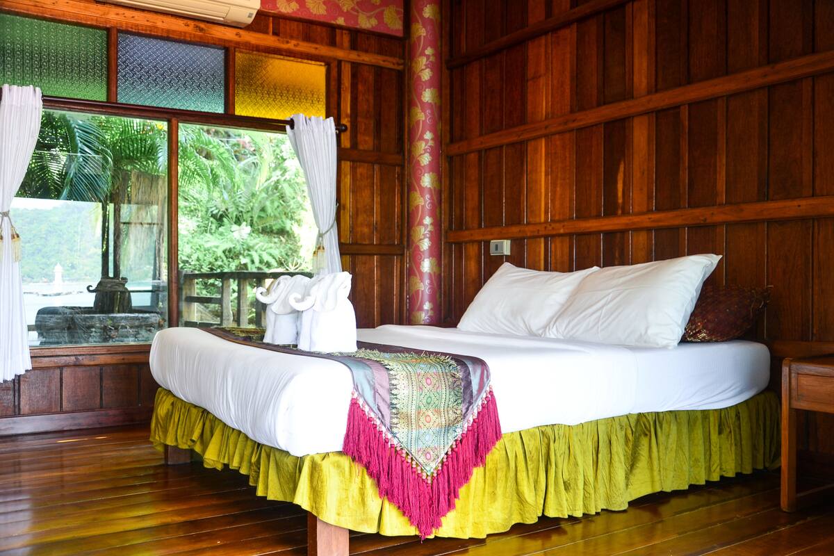 A wooden room with a bed, a window with curtains, and a view of greenery outside.