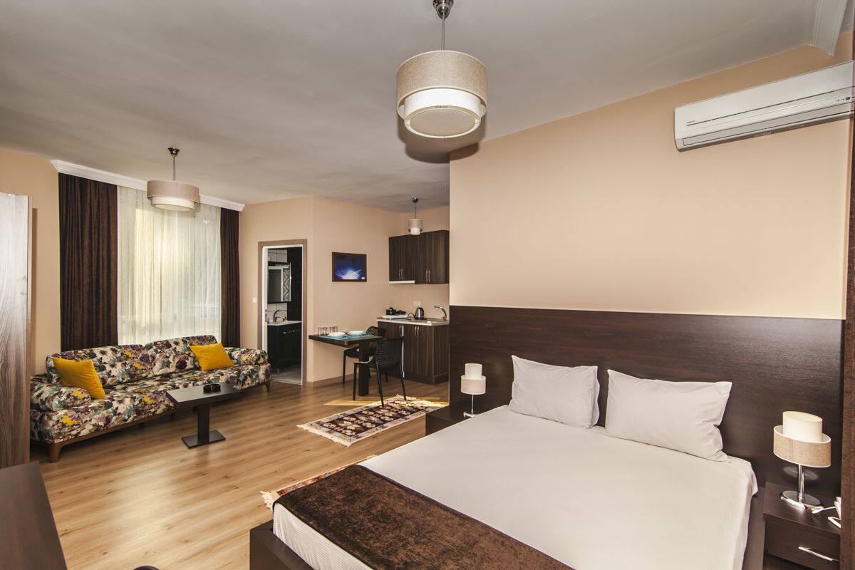 A hotel room with a large bed, a sofa with yellow cushions, a desk with a chair, and a kitchen area.