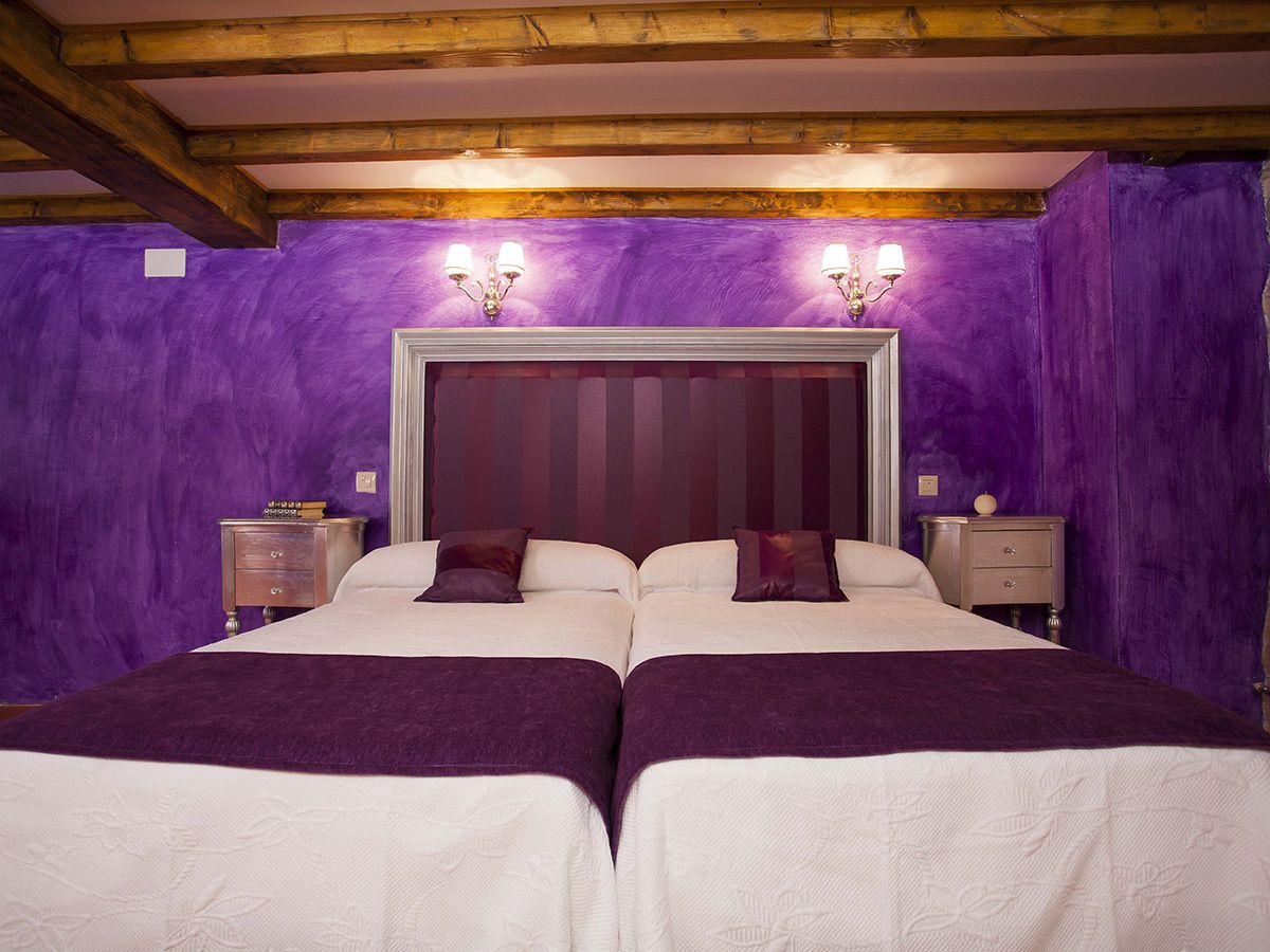 Een tweepersoonskamer met twee bedden, paarse muren, houten balken en bedlampjes.