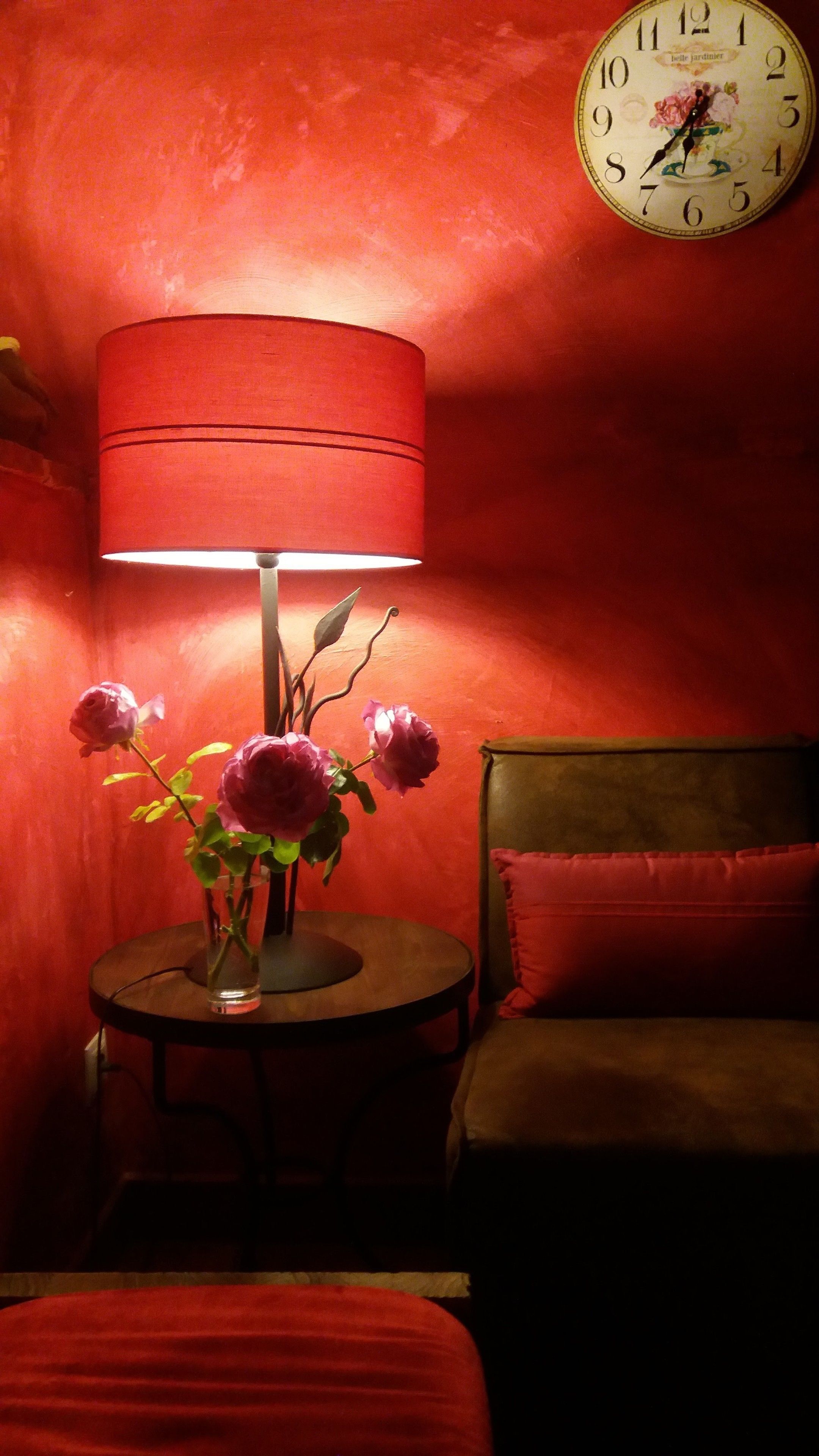 A room with a round table, a red lamp, a vase with flowers, a clock, and a bed with red bedding.