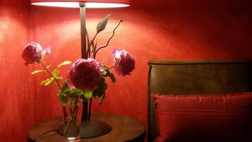 A room with a round table, a red lamp, a vase with flowers, a clock, and a bed with red bedding.