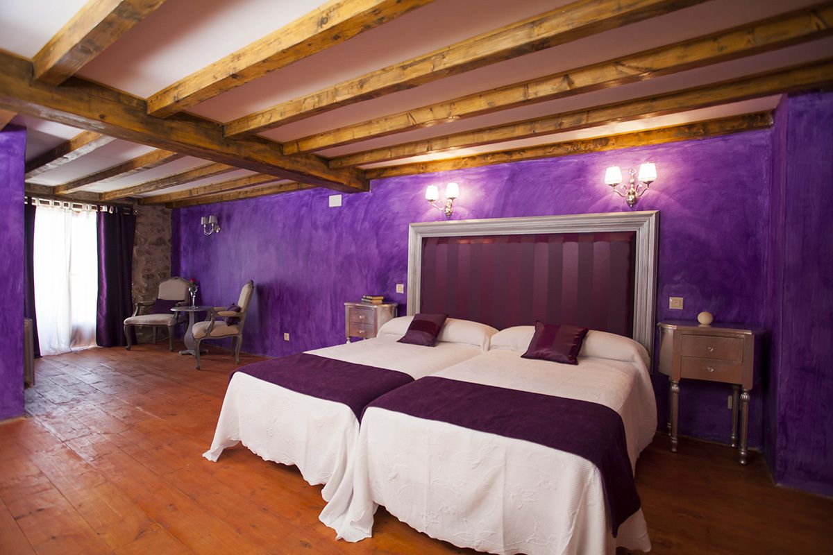 A room with two beds, wooden beams, purple walls, and a stone wall.