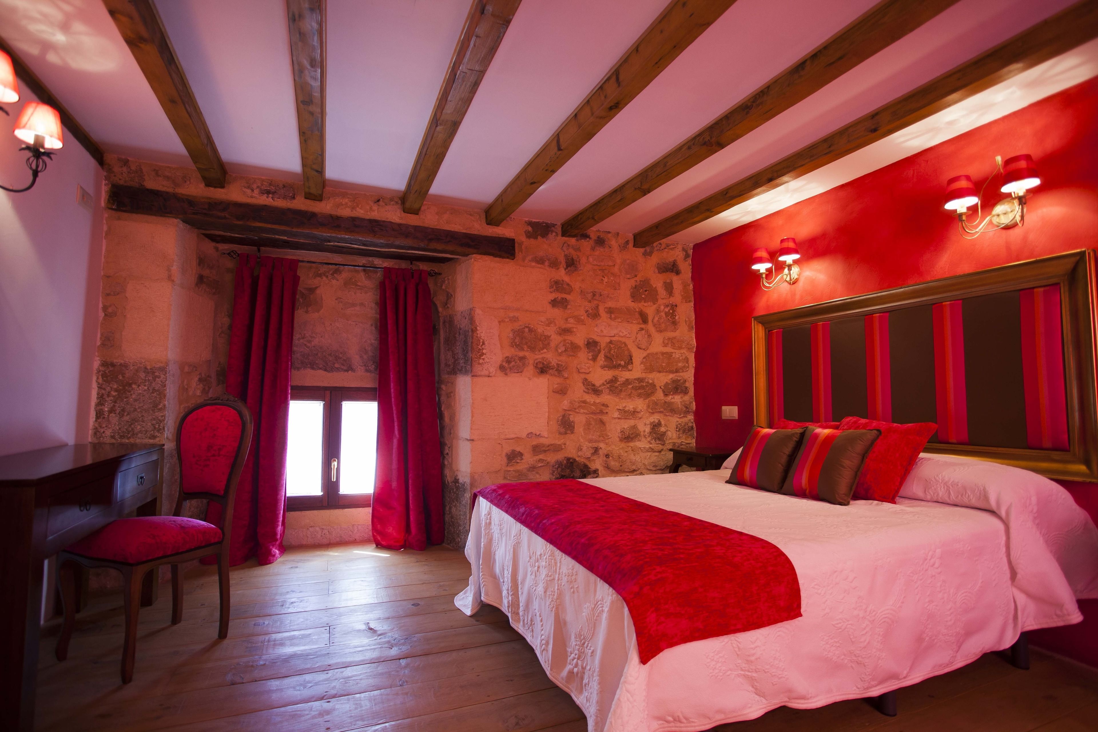 Un dormitorio con pared de piedra, vigas de madera, una cama con ropa de cama roja, un escritorio y una silla.