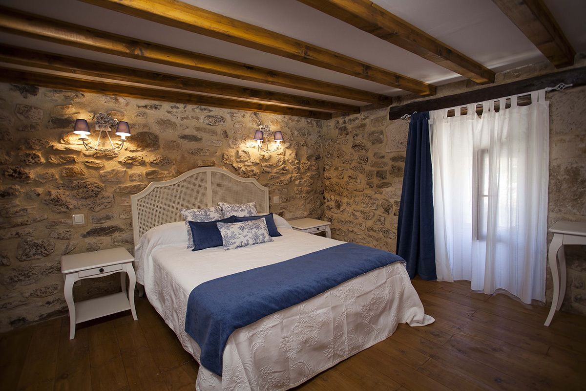 Un dormitorio con pared de piedra, vigas de madera, una cama con cabecera blanca, dos mesitas de noche y una ventana con cortinas.