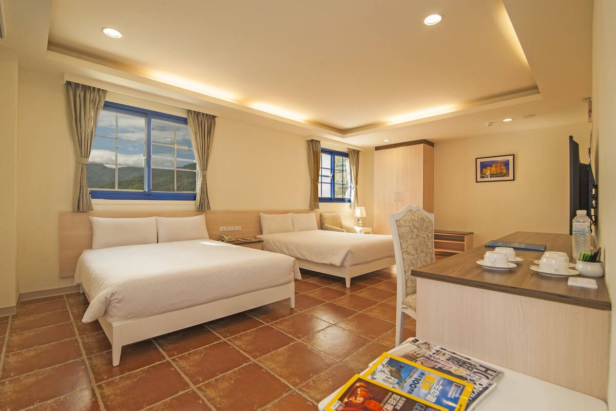 A hotel room with two beds, a desk, a chair, a window with curtains, a painting on the wall, and a tiled floor.