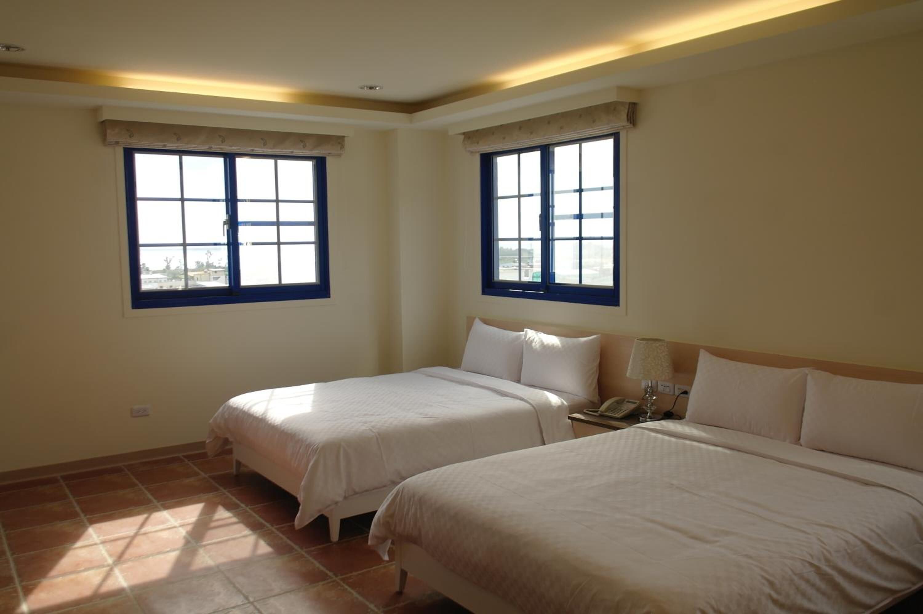 Ein Hotelzimmer mit zwei Betten, einem Nachttisch mit Lampe und zwei Fenstern mit Vorhängen.