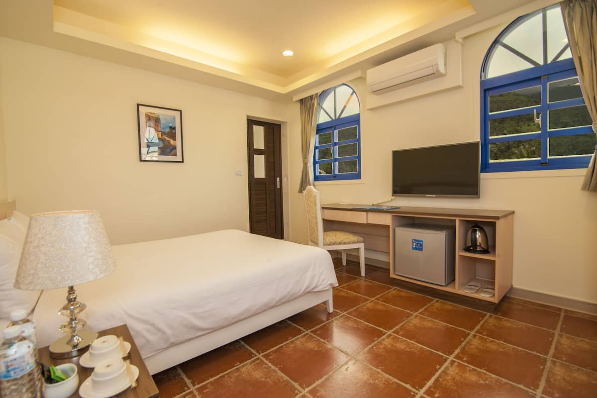 A hotel room with a bed, a desk with a television, a small refrigerator, and a window with curtains.