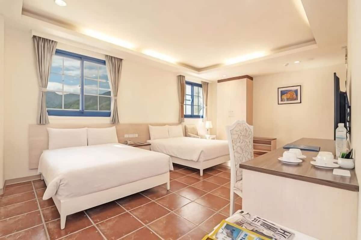 A hotel room with two beds, a desk, a chair, a window with curtains, and a painting on the wall.