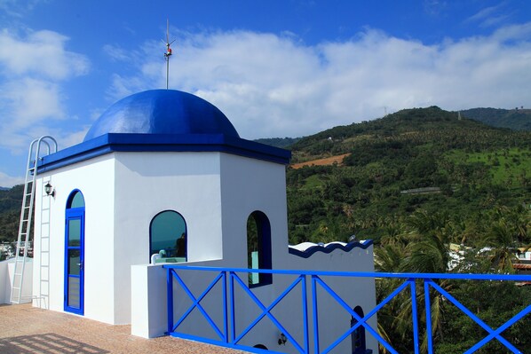 A white building with a blue dome and arched windows, surrounded by lush green hills and a clear blue sky.