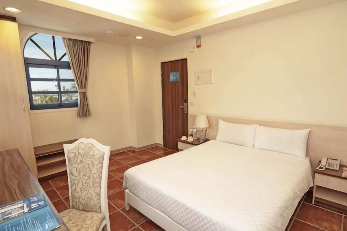 A hotel room with a large bed, a desk, a chair, a window with curtains, and a door.