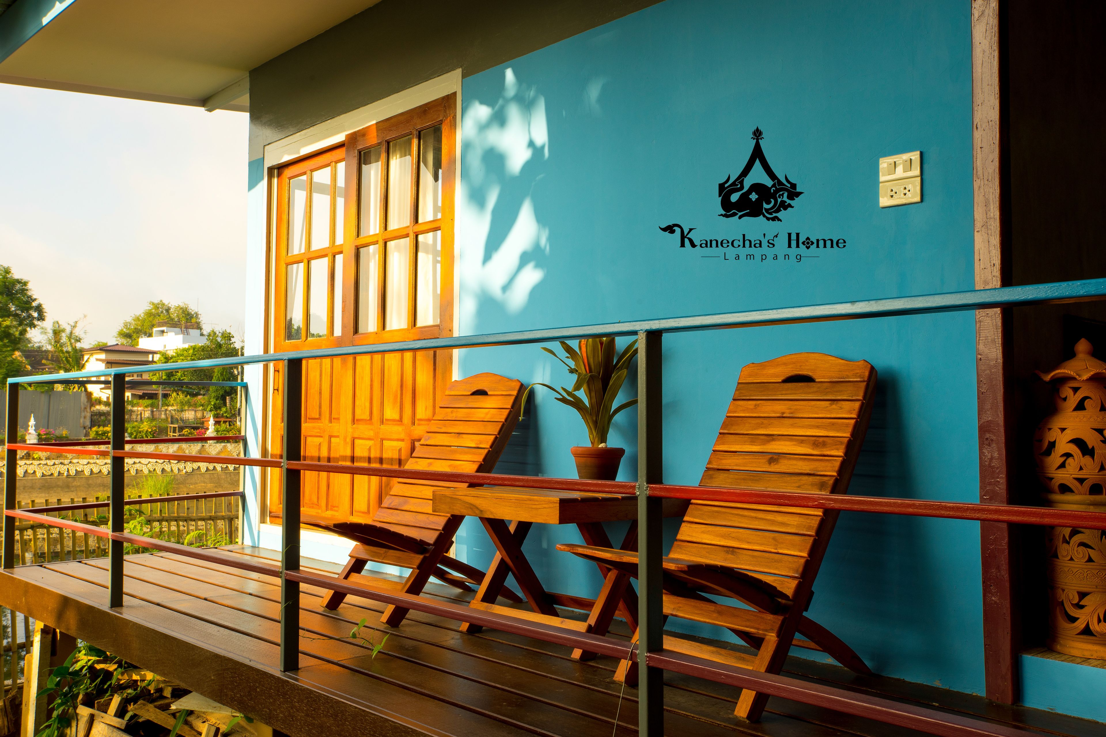A wooden deck with two lounge chairs and a potted plant, a blue wall with a logo, and a window with curtains.