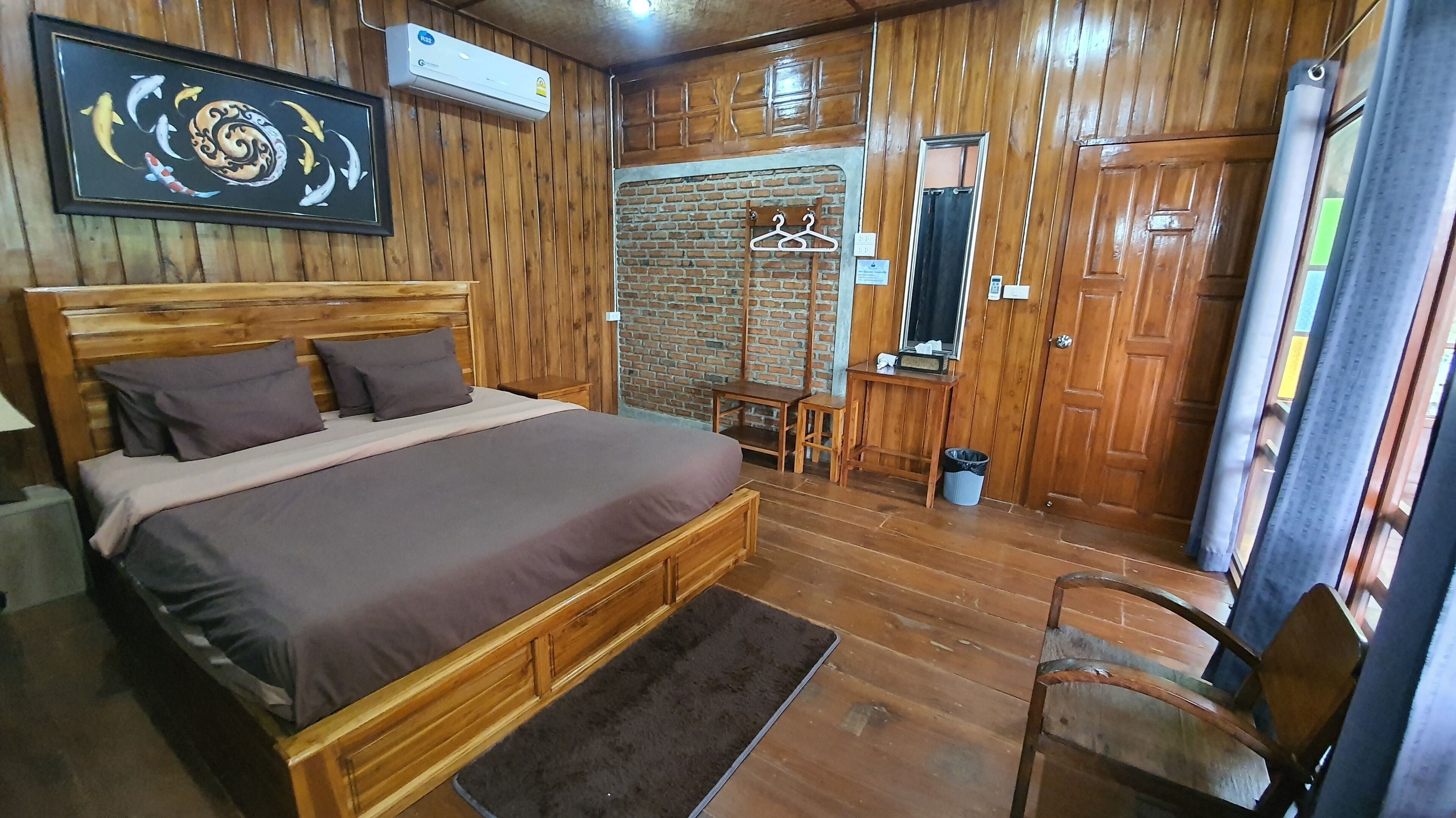 A wooden-furnished bedroom with a bed, a wooden headboard, a wall-mounted air conditioner, a framed picture, a wooden door, and a small table with a chair.