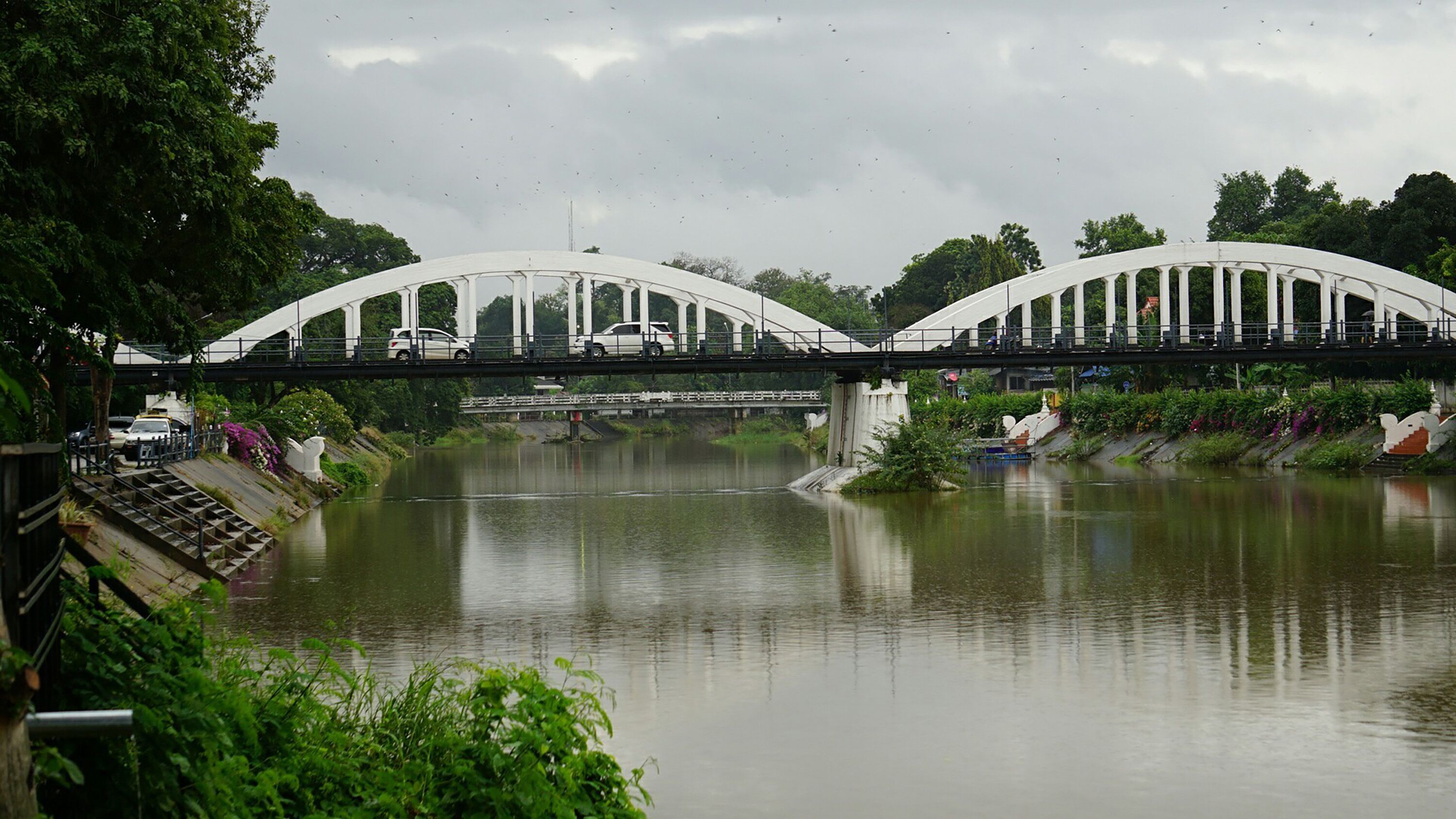 A white bridge with a curved design spanning over a river, surrounded by greenery.