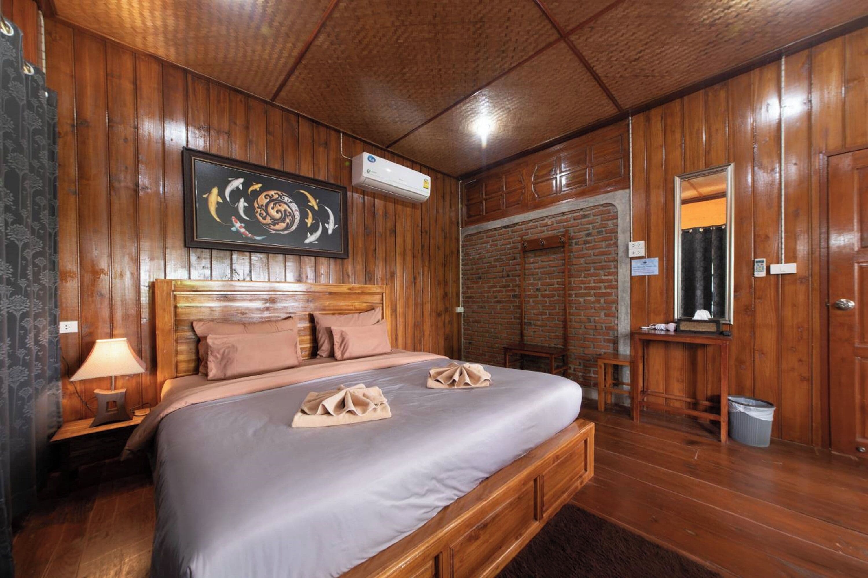A wooden-paneled room with a bed, a nightstand, a lamp, a framed artwork, a brick wall, and a mirror.