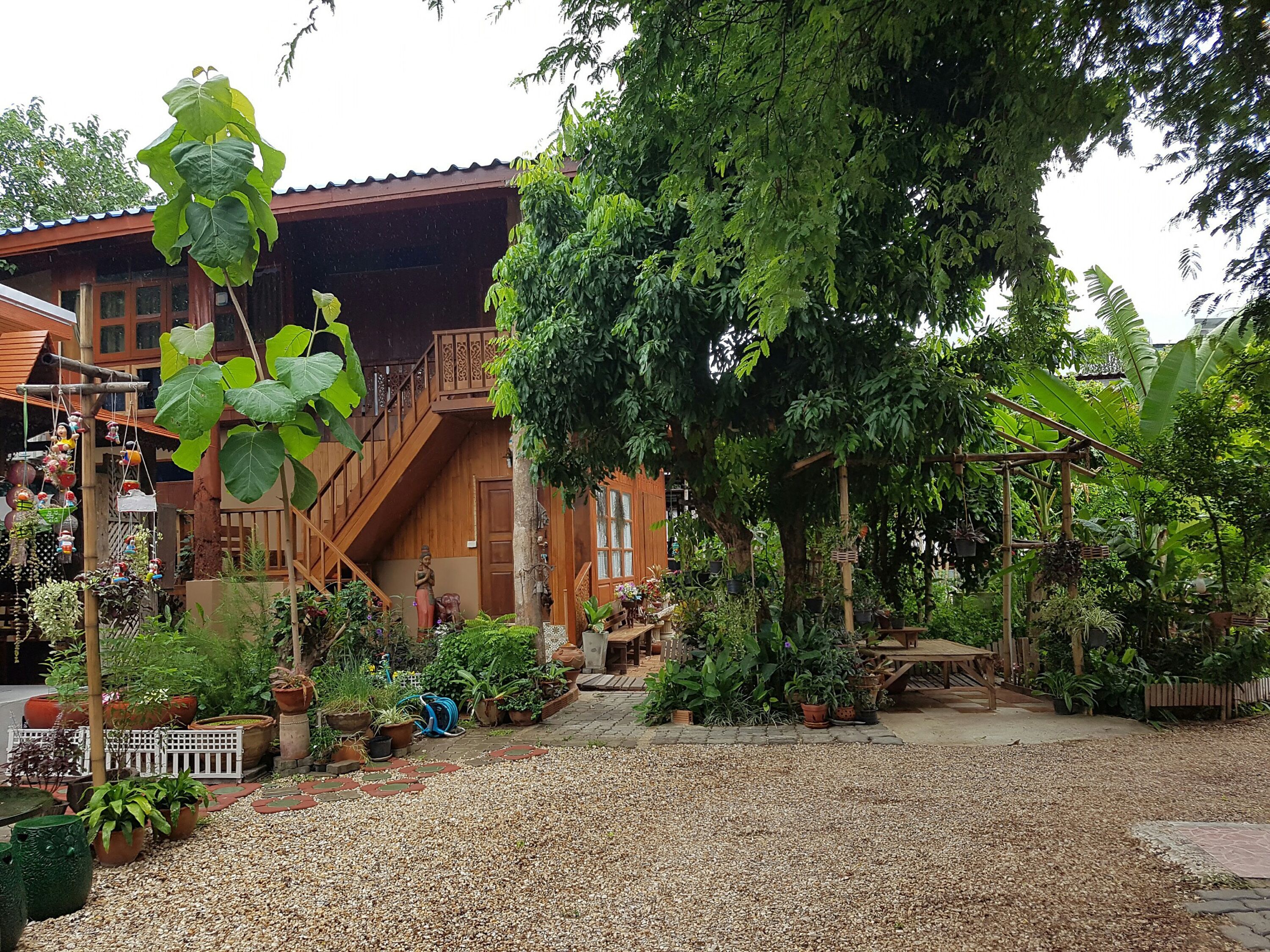 Une maison en bois avec un escalier menant à un balcon, entourée de plantes en pot et d’un chemin de gravier.