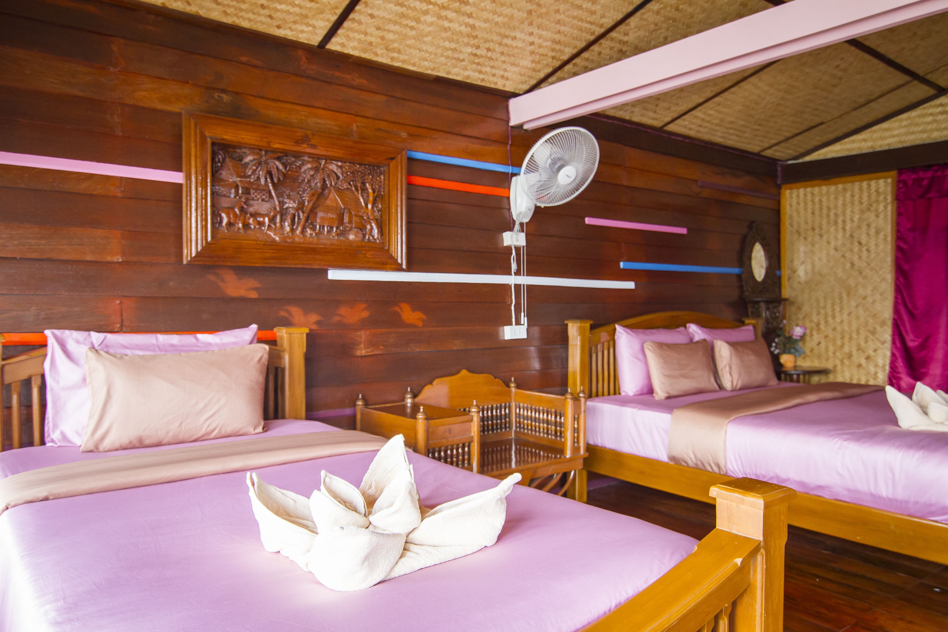A room with two beds, each with a wooden headboard, a bedside table, and a wall-mounted fan.