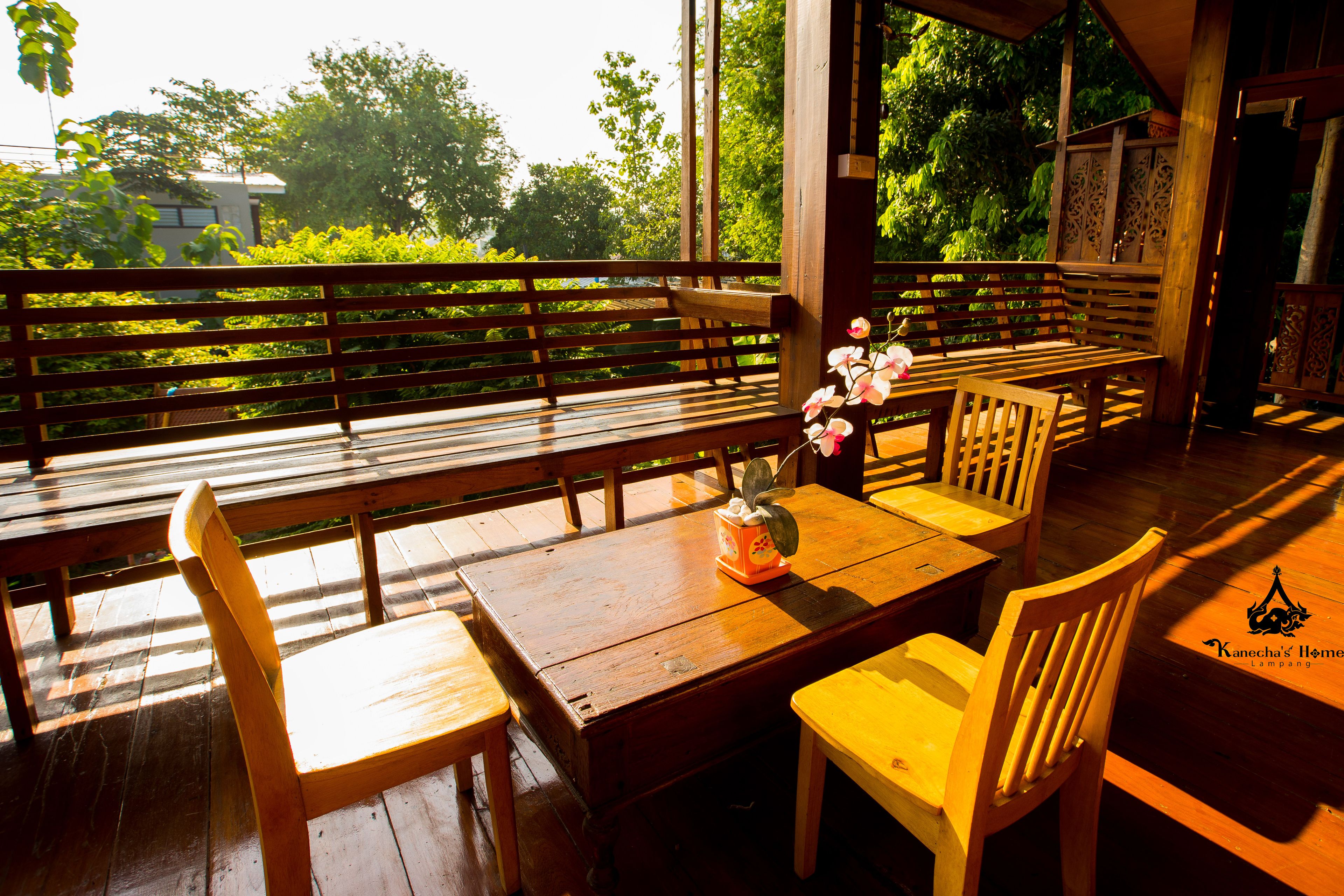 A wooden deck with benches and chairs, a table with a vase of flowers, and a view of greenery and trees.