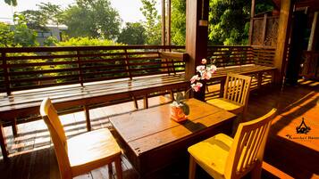 A wooden deck with benches and chairs, a table with a vase of flowers, and a view of greenery and trees.