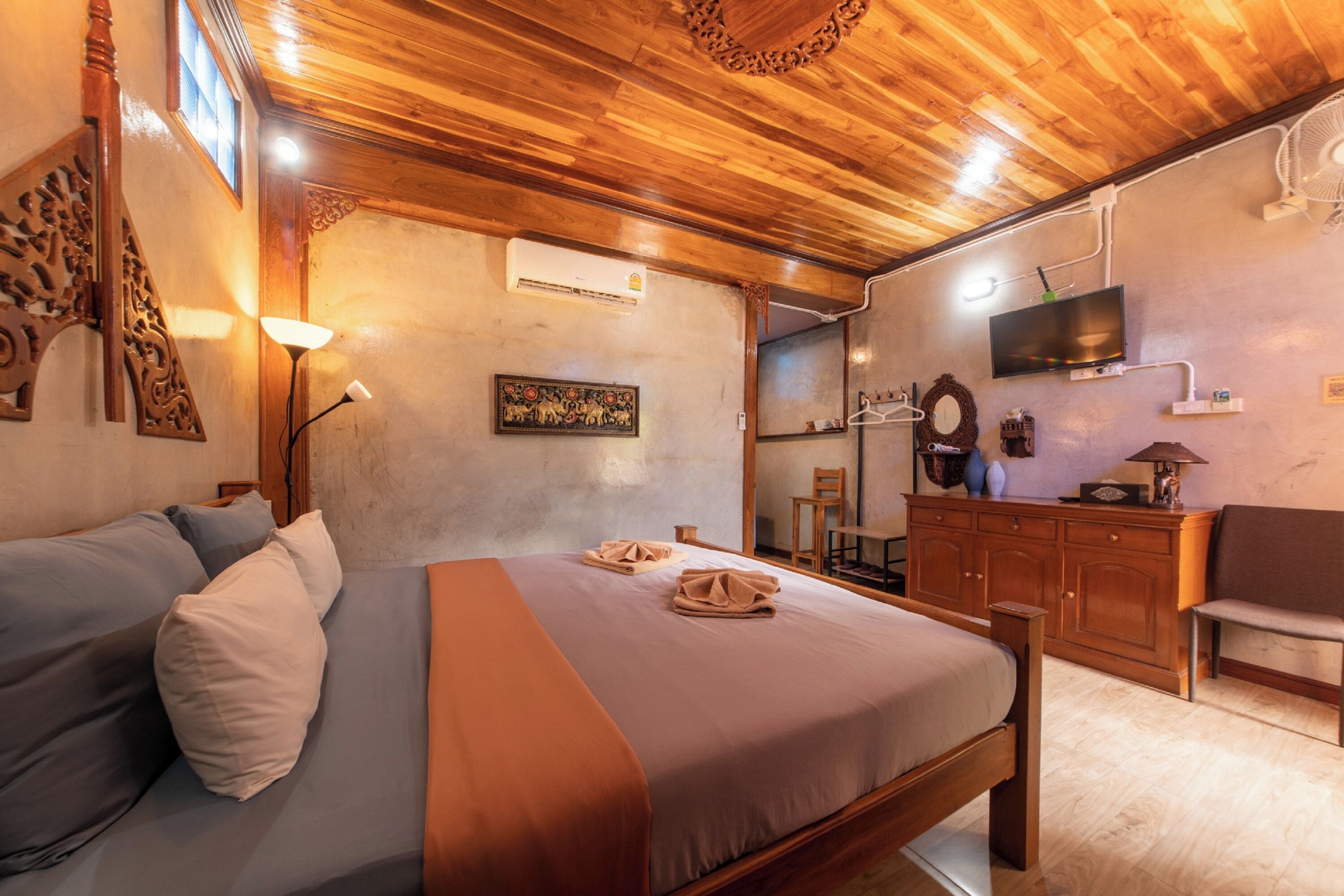A bedroom with a wooden ceiling, a bed with a brown bedspread, a wooden dresser, a television, and a mirror.
