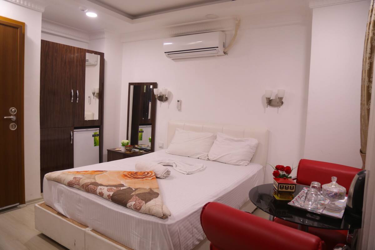 Ein Hotelzimmer mit einem Bett, einem roten Sessel, einem kleinen Tisch mit einer Uhr und Dekoration, einer Klimaanlage und einem Kleiderschrank.