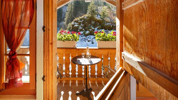 A wooden balcony with a table and chairs, a view of snow-covered trees, and a window with curtains. - Griesalp Hotels (Reichenbach im Kandertal)