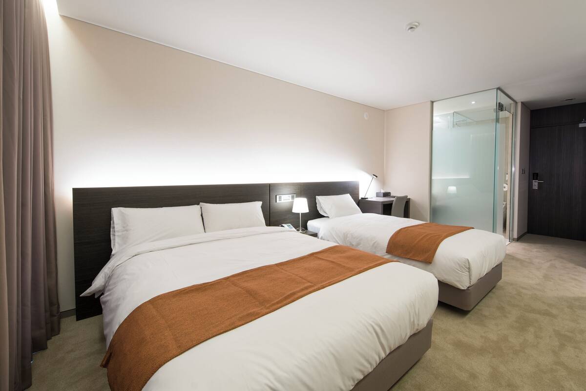 A hotel room with two beds, a desk, a chair, a nightstand, and a glass door leading to a bathroom.