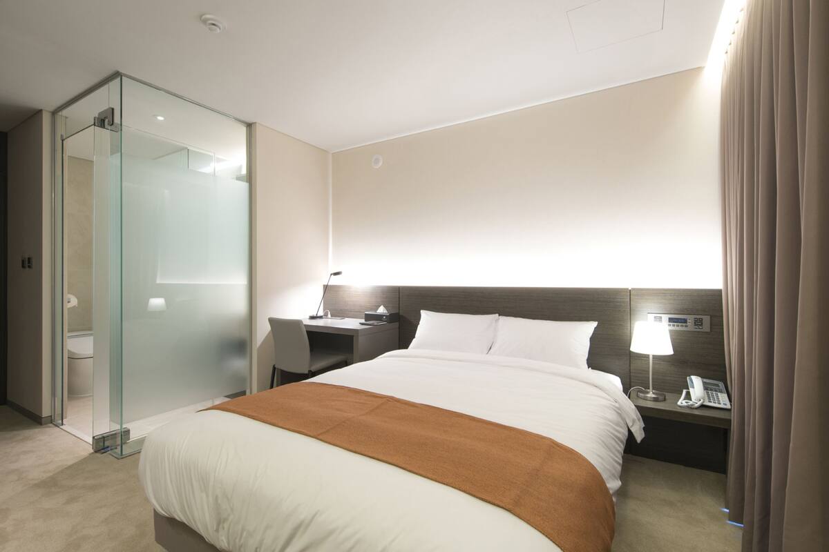 A hotel room with a bed, a desk, a chair, a glass shower enclosure, and a nightstand with a lamp and phone.